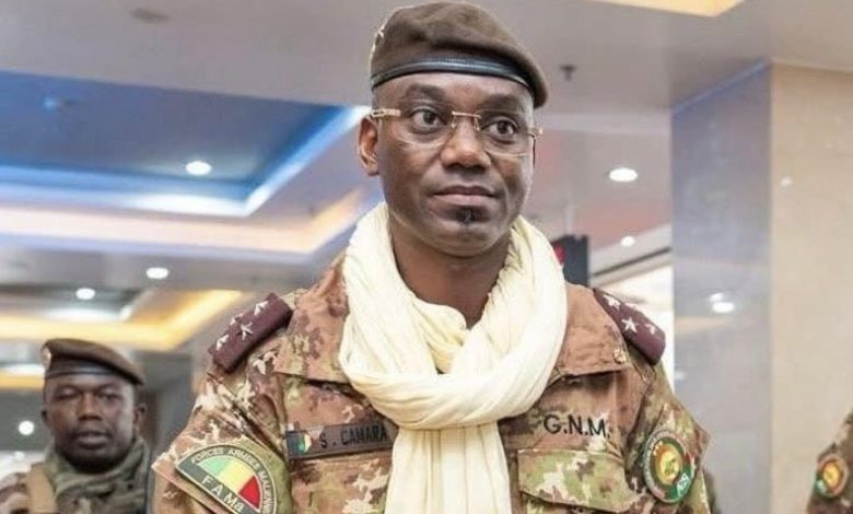 Man in camouflage military uniform and beret, wearing a light scarf, standing in an indoor hallway.”