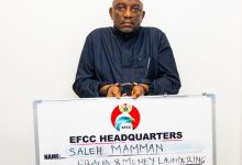 Handcuffed man in dark traditional clothing stands behind an EFCC Headquarters signboard showing charges, including fraud and money laundering.