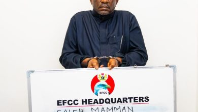 Handcuffed man in dark traditional clothing stands behind an EFCC Headquarters signboard showing charges, including fraud and money laundering.