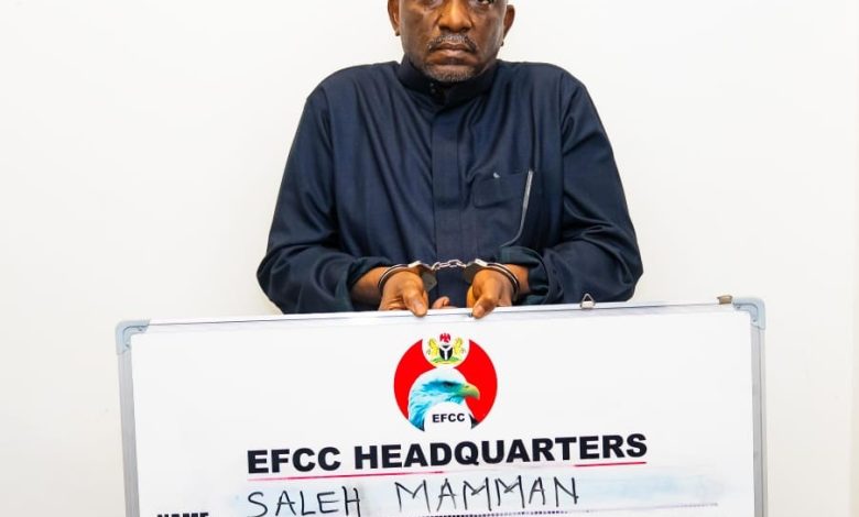 Saleh Momoh - The Times of Abuja Handcuffed man in dark traditional clothing stands behind an EFCC Headquarters signboard showing charges, including fraud and money laundering.
