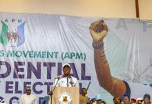 Speaker at a podium addressing a crowd on a stage with a large banner behind him showing the raised fist graphic and 'APM' lettering