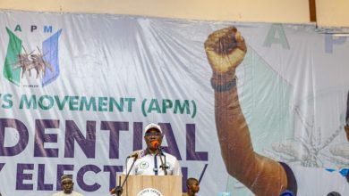Speaker at a podium addressing a crowd on a stage with a large banner behind him showing the raised fist graphic and 'APM' lettering
