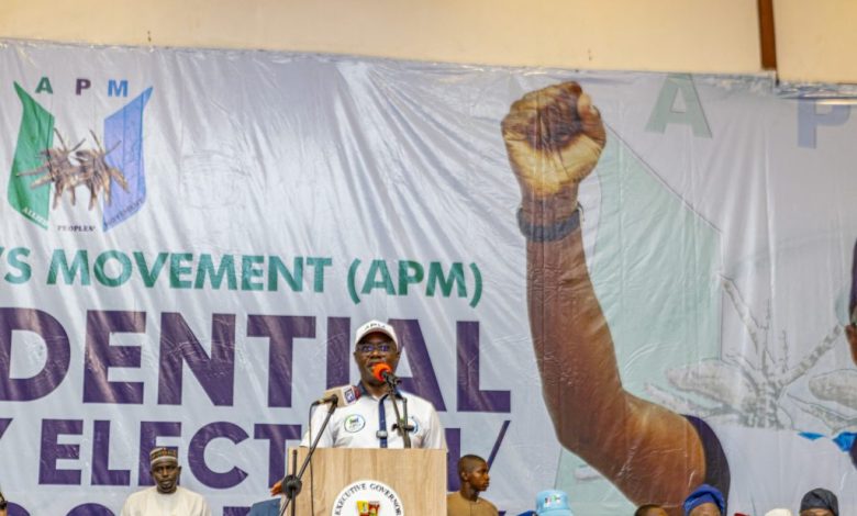 Seyi Makinde 1 - The Times of Abuja Speaker at a podium addressing a crowd on a stage with a large banner behind him showing the raised fist graphic and 'APM' lettering