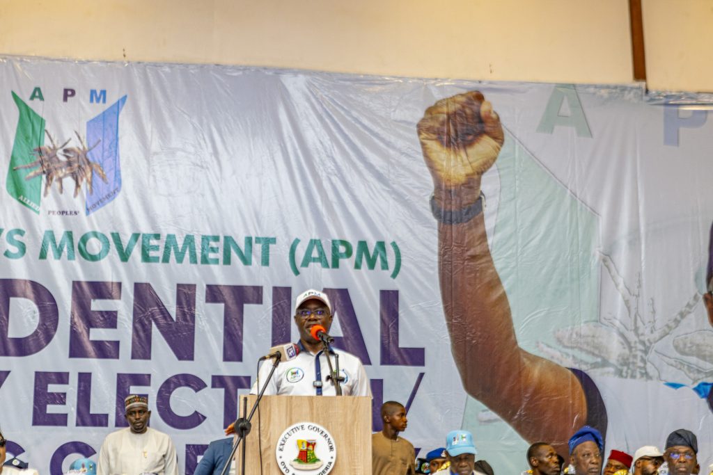 Seyi Makinde 1 - The Times of Abuja Speaker at a podium addressing a crowd on a stage with a large banner behind him showing the raised fist graphic and'APM' lettering