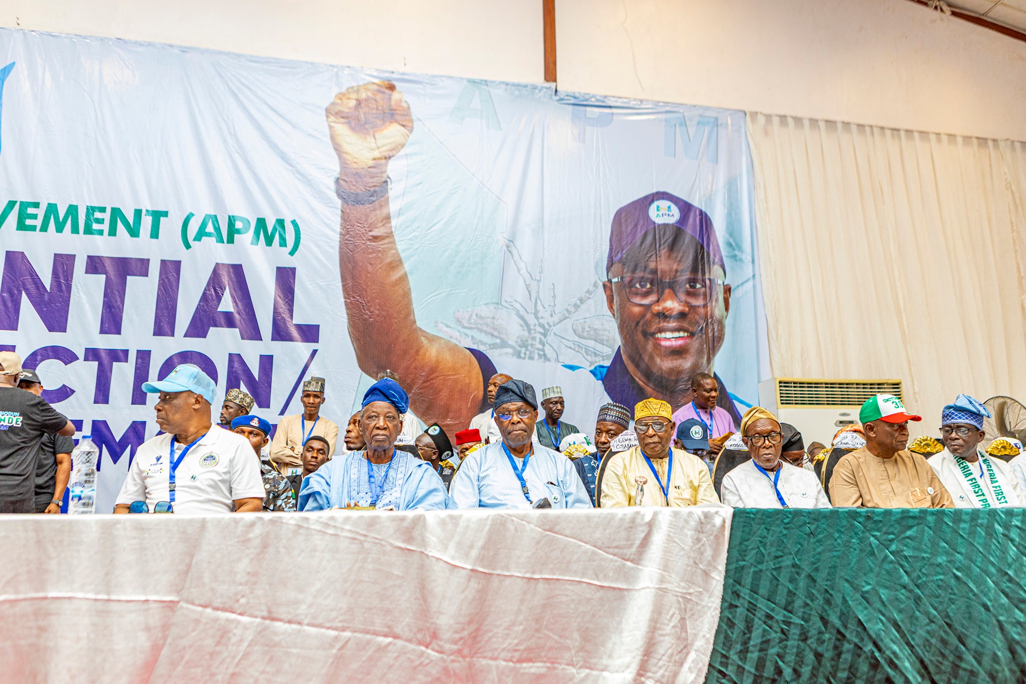 Seyi Makinde 3 - The Times of Abuja Group of people seated on a stage at a political event in front of a large banner featuring a raised fist and the letters AP M/NG (APM).