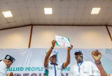 People on a stage raise a certificate in celebration, smiling as others applaud in the background, with a banner behind them reading Allied People’s Movement.