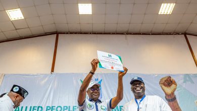 People on a stage raise a certificate in celebration, smiling as others applaud in the background, with a banner behind them reading Allied People’s Movement.