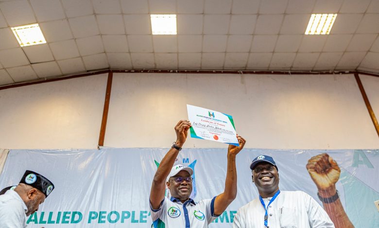 Seyi Makinde 4 - The Times of Abuja People on a stage raise a certificate in celebration, smiling as others applaud in the background, with a banner behind them reading Allied People’s Movement.