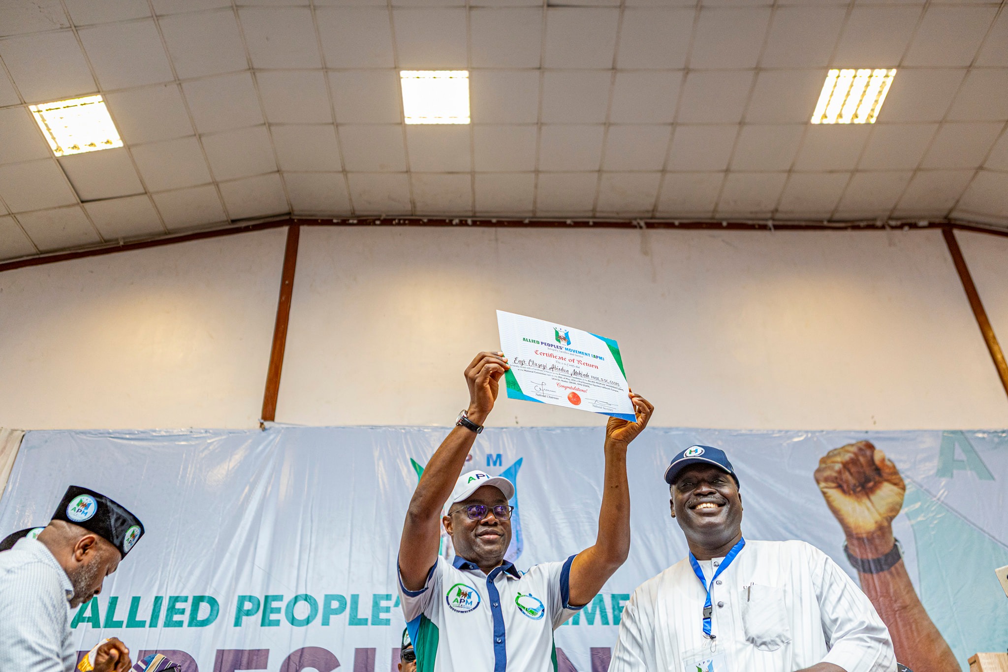 Seyi Makinde 4 - The Times of Abuja People on a stage raise a certificate in celebration, smiling as others applaud in the background, with a banner behind them reading Allied People’s Movement.