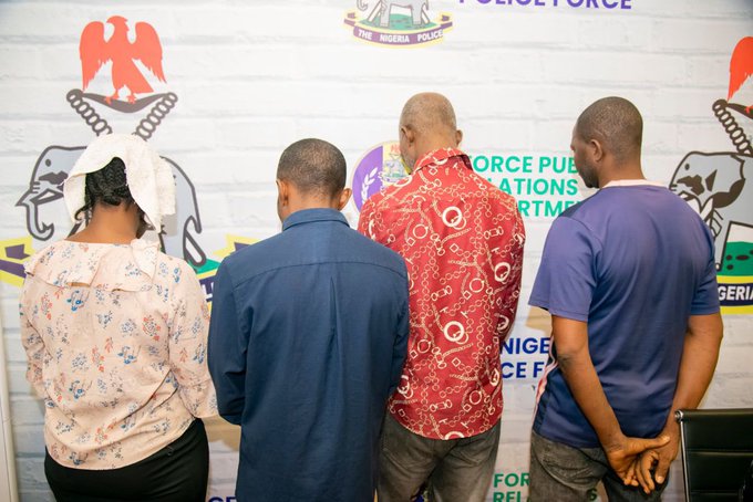 Four men stand with their backs to the camera in a police lineup against a wall with logos and text behind them (front-facing away).