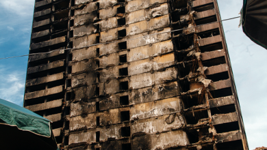 Charred, fire-damaged high-rise facade with exposed rebar and burnt concrete under a blue sky.