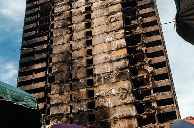 Architecture as Wound - The Times of Abuja Charred, fire-damaged high-rise facade with exposed rebar and burnt concrete under a blue sky.