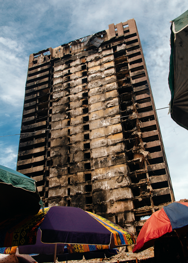 Architecture as Wound - The Times of Abuja Tall burned-out apartment building with a charred façade rising over a busy outdoor market with colorful umbrellas.