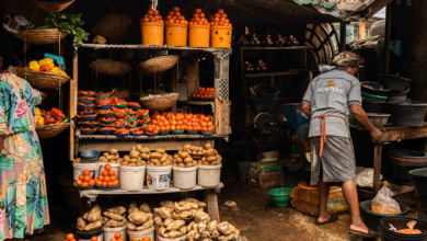 Outdoor market stall with baskets of tomatoes, ginger, and potatoes on display; a vendor in a gray apron works behind the counter.