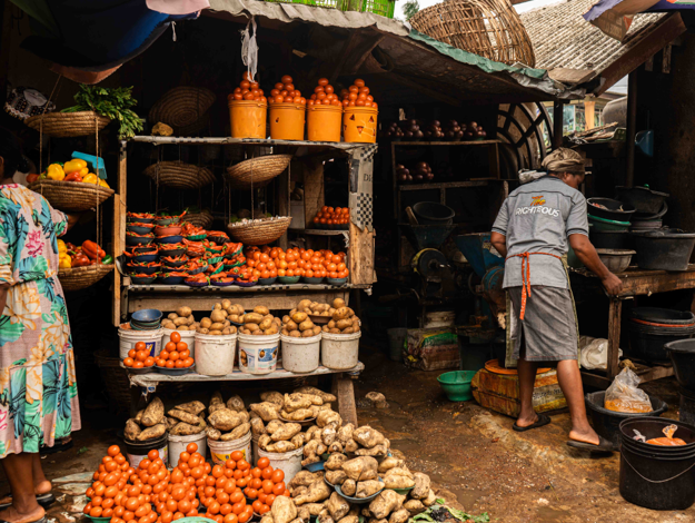 Colour of Abundance - The Times of Abuja Outdoor market stall with baskets of tomatoes, ginger, and potatoes on display; a vendor in a gray apron works behind the counter.