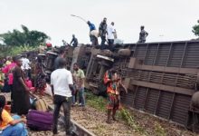 Crowd gathers around a derailed freight train lying on its side with people standing on and near the overturned cars on the tracks
