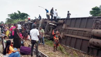 Crowd gathers around a derailed freight train lying on its side with people standing on and near the overturned cars on the tracks