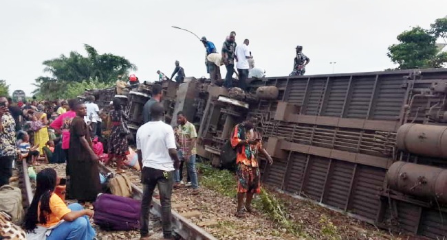 Crowd gathers around a derailed freight train lying on its side with people standing on and near the overturned cars on the tracks