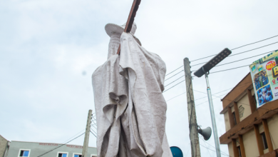 Statue wrapped in white fabric on a city street, with buildings, utility poles, and a billboard in the background.