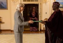 Woman in a gray suit hands a green folder to a man in ceremonial robes as another woman watches from the doorway.