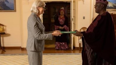 Woman in a gray suit hands a green folder to a man in ceremonial robes as another woman watches from the doorway.