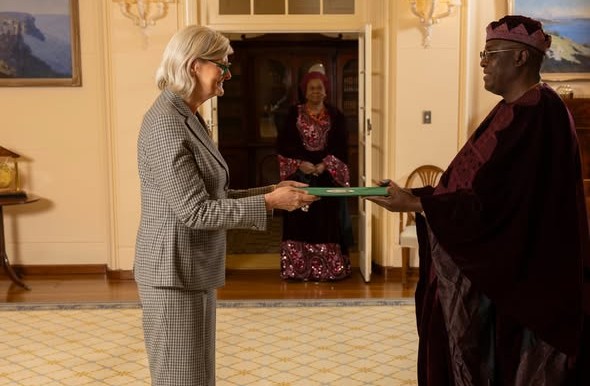 Femi Pedro - The Times of Abuja Woman in a gray suit hands a green folder to a man in ceremonial robes as another woman watches from the doorway.