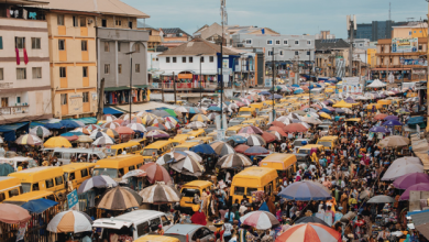Crowded market street filled with colorful umbrellas shading stalls, pedestrians, and yellow minibuses lining the road.
