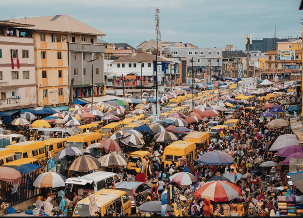 Crowded market street filled with colorful umbrellas shading stalls, pedestrians, and yellow minibuses lining the road.