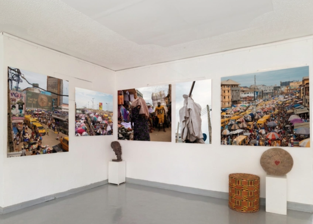 Interior of a small art gallery with five large market-scene photos on white walls, and sculptures on pedestals plus a woven basket and carved heart nearby.