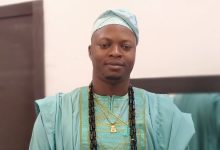 Man wearing turquoise traditional African agbada and cap with long beaded necklaces, facing forward in a formal portrait.