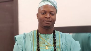 Man wearing turquoise traditional African agbada and cap with long beaded necklaces, facing forward in a formal portrait.