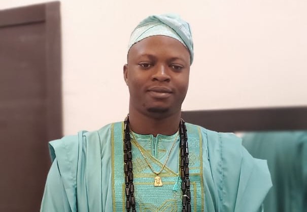 Man wearing turquoise traditional African agbada and cap with long beaded necklaces, facing forward in a formal portrait.