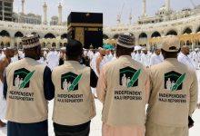 Four reporters wearing beige vests with an 'Independent Hajj Reporters' logo stand with their backs to the camera near the Kaaba in Mecca during Hajj.