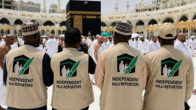 Four reporters wearing beige vests with an 'Independent Hajj Reporters' logo stand with their backs to the camera near the Kaaba in Mecca during Hajj.
