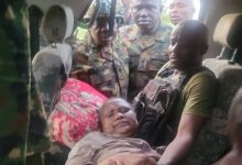 Woman lying on a car seat as soldiers surround her and assist, with a man supporting her on the right.