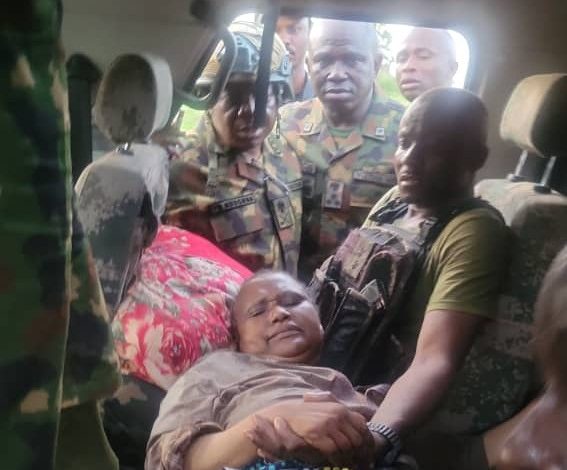Woman lying on a car seat as soldiers surround her and assist, with a man supporting her on the right.