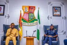 Two men seated in ornate armchairs in a formal government room; one in tan traditional attire speaks into a microphone, the other in a blue military uniform sits with notes, with a crest, flags, and microphones between them.