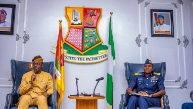 Two men seated in ornate armchairs in a formal government room; one in tan traditional attire speaks into a microphone, the other in a blue military uniform sits with notes, with a crest, flags, and microphones between them.