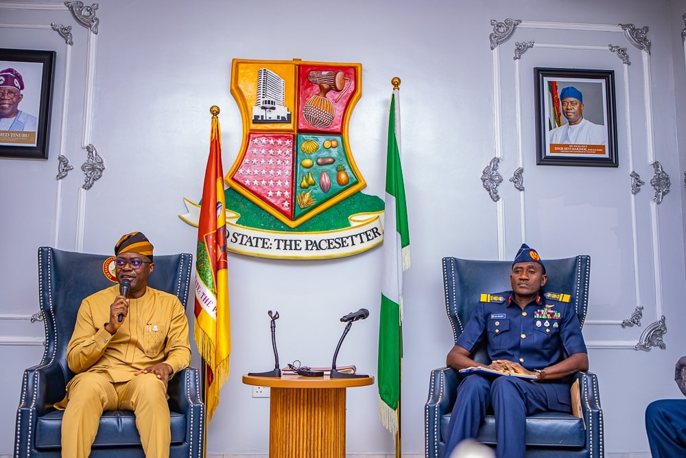 Makinde and NAF officers 2 - The Times of Abuja Two men seated in ornate armchairs in a formal government room; one in tan traditional attire speaks into a microphone, the other in a blue military uniform sits with notes, with a crest, flags, and microphones between them.