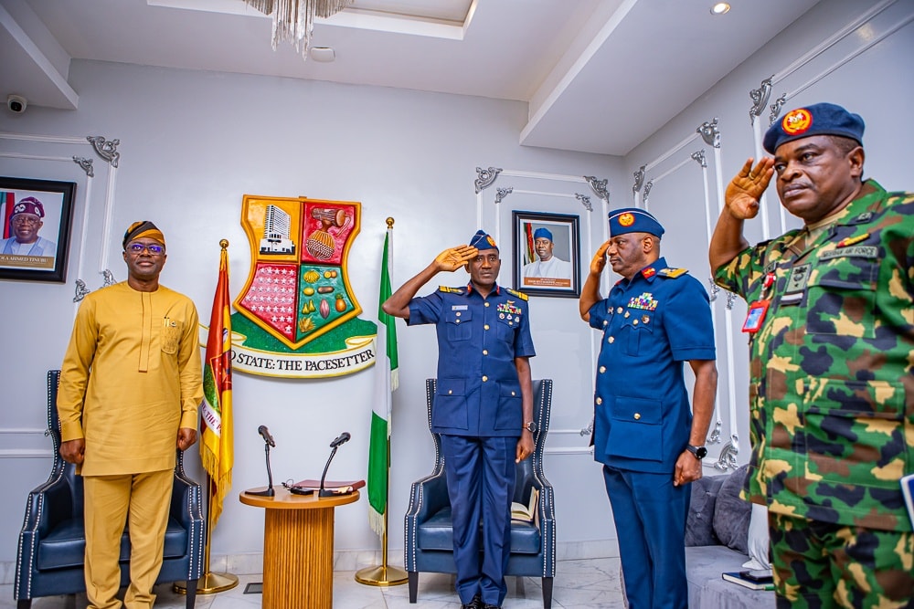 Makinde and NAF officers 5 - The Times of Abuja Five men stand in a formal room, saluting: one in tan traditional attire and four in blue military uniforms,with a coat of arms, flags, and portraits behind them.