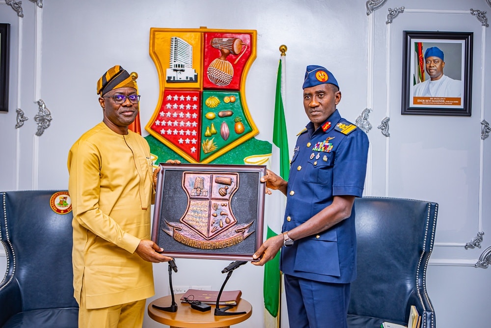 Makinde and NAF officers - The Times of Abuja Two men in formal attire exchange a framed commemorative plaque during a ceremony in a government office, with a crest on the wall behind them.