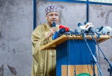 Male official in a beige traditional outfit speaks at a podium with many microphones during a press conference; Nigerian flag on the left.