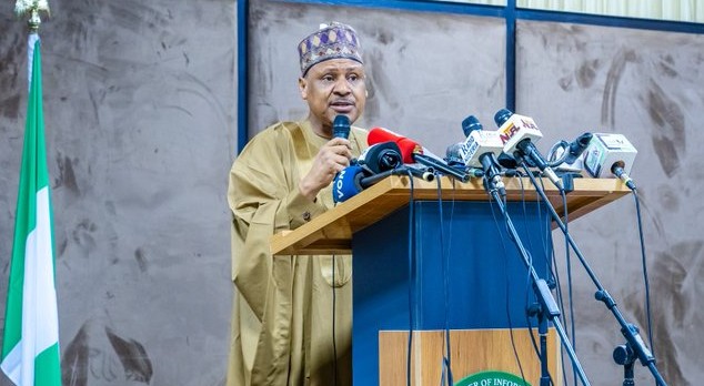 Male official in a beige traditional outfit speaks at a podium with many microphones during a press conference; Nigerian flag on the left.