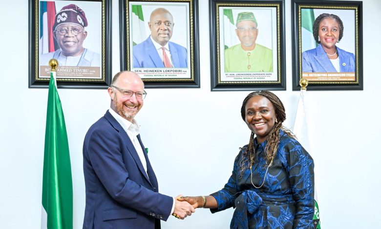 NURPC 1 - The Times of Abuja Two people shake hands in a formal setting with framed official portraits on the wall behind them and green‑white flags visible on either side.