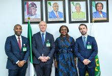 Four professionally dressed individuals stand side by side in front of framed portraits on a wall, with a Nigerian flag nearby.