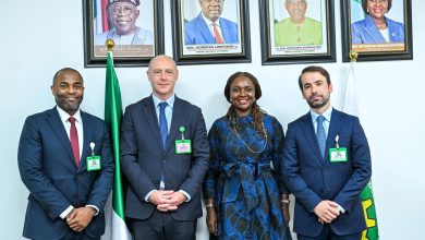 Four professionally dressed individuals stand side by side in front of framed portraits on a wall, with a Nigerian flag nearby.