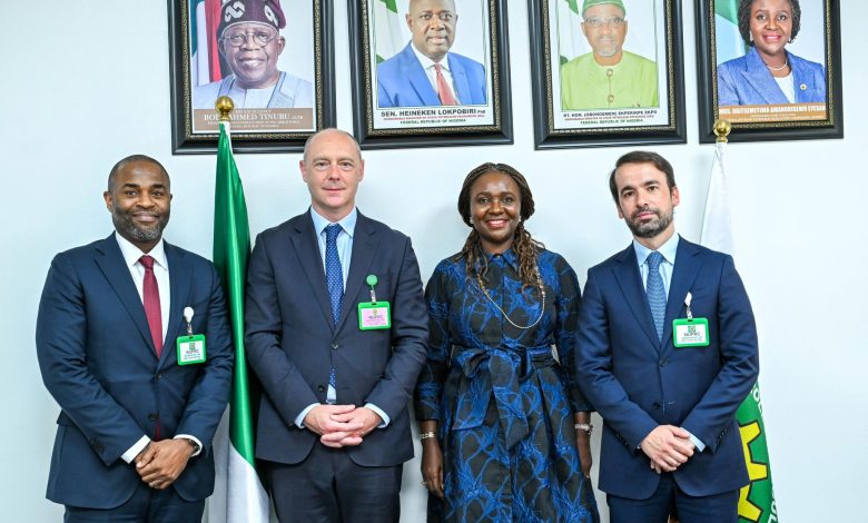 Four professionally dressed individuals stand side by side in front of framed portraits on a wall, with a Nigerian flag nearby.