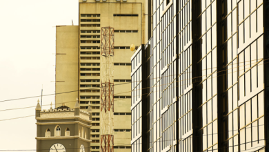 Urban street scene with a tall beige building marked 'NAI', a clock tower, and a glass-fronted office building on the right.