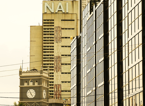No Time - The Times of Abuja Urban street scene with a tall beige building marked 'NAI', a clock tower, and a glass-fronted office building on the right.
