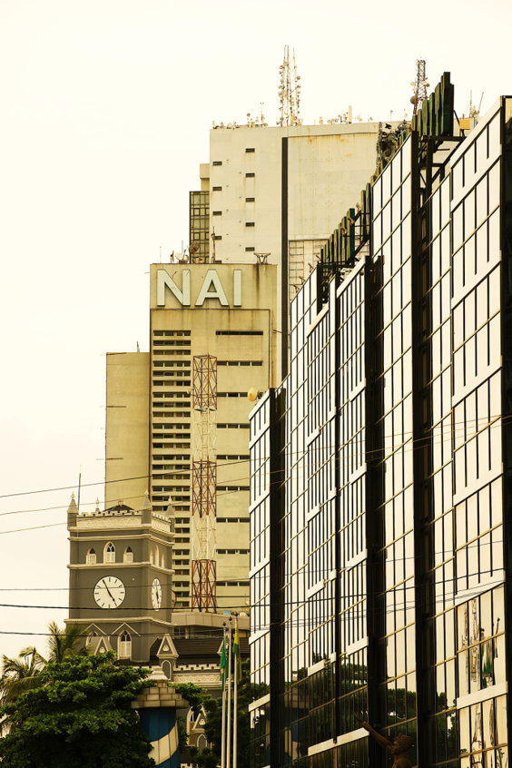 No Time - The Times of Abuja Urban street scene with a beige NAI building, a clock tower, and a tall glass-fronted office on a pale sky backdrop